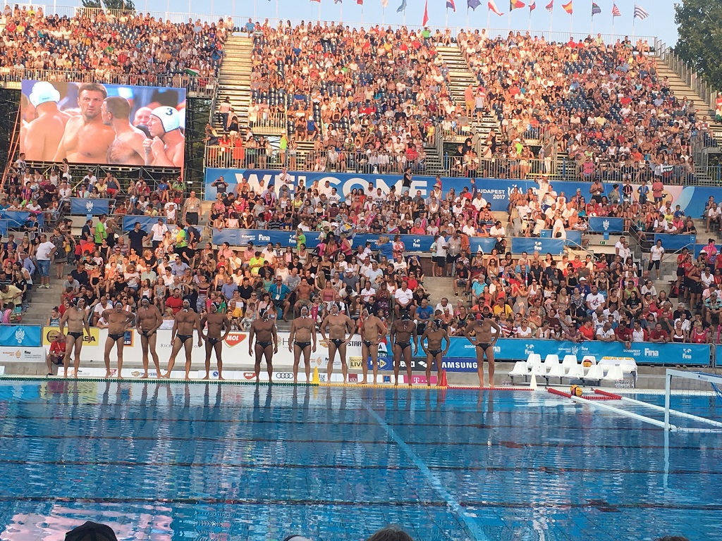 Darkside Water Polo team lined up by the pool at the 2017 Budapest Championships, with a large crowd in the stands and international flags above.